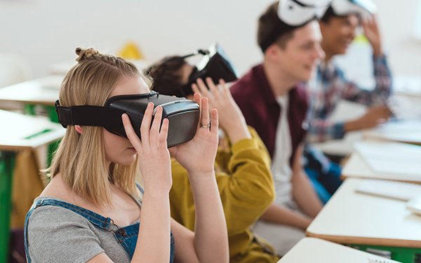 Zu sehen sind, hinter Schulbänken sitzende, Jugendliche, die durch VR- Brillen schauen.