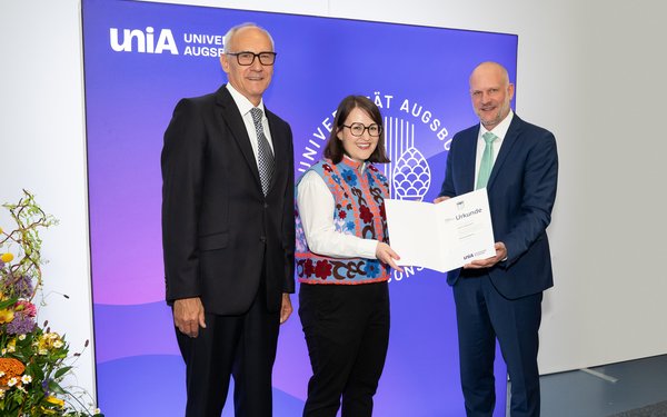 Drei Personen, in der Mitte steht Dr. Katja Boser, der ein Dokument (die Urkunde für den Wissenschaftspreis) verliehen wird