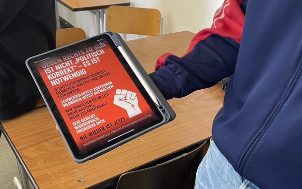 Ein Schüler h?lt ein Tablet in die H?he. Darauf ist auf rotem Hintergrund wei?e und schwarze Schrift und eine wei?e Faust zu sehen.