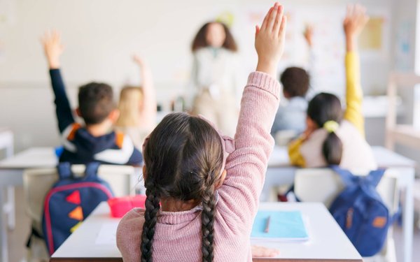 Eine Schulklasse von hinten. Ein Mädchen mit dunklen Zöpfen im Vordergrund des Bildes streckt die Hand. Die Lehrerin vorne ist nur verschwommen zu sehen.