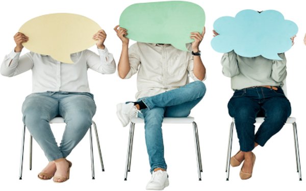 Vier Personen sitzen nebeneinander auf St��hlen. Die Personen tragen alle Jeanshosen und ein helles Oberteil. Die Personen halten sich bunte Schilder in Form von Sprechblasen vor ihre Gesichter.