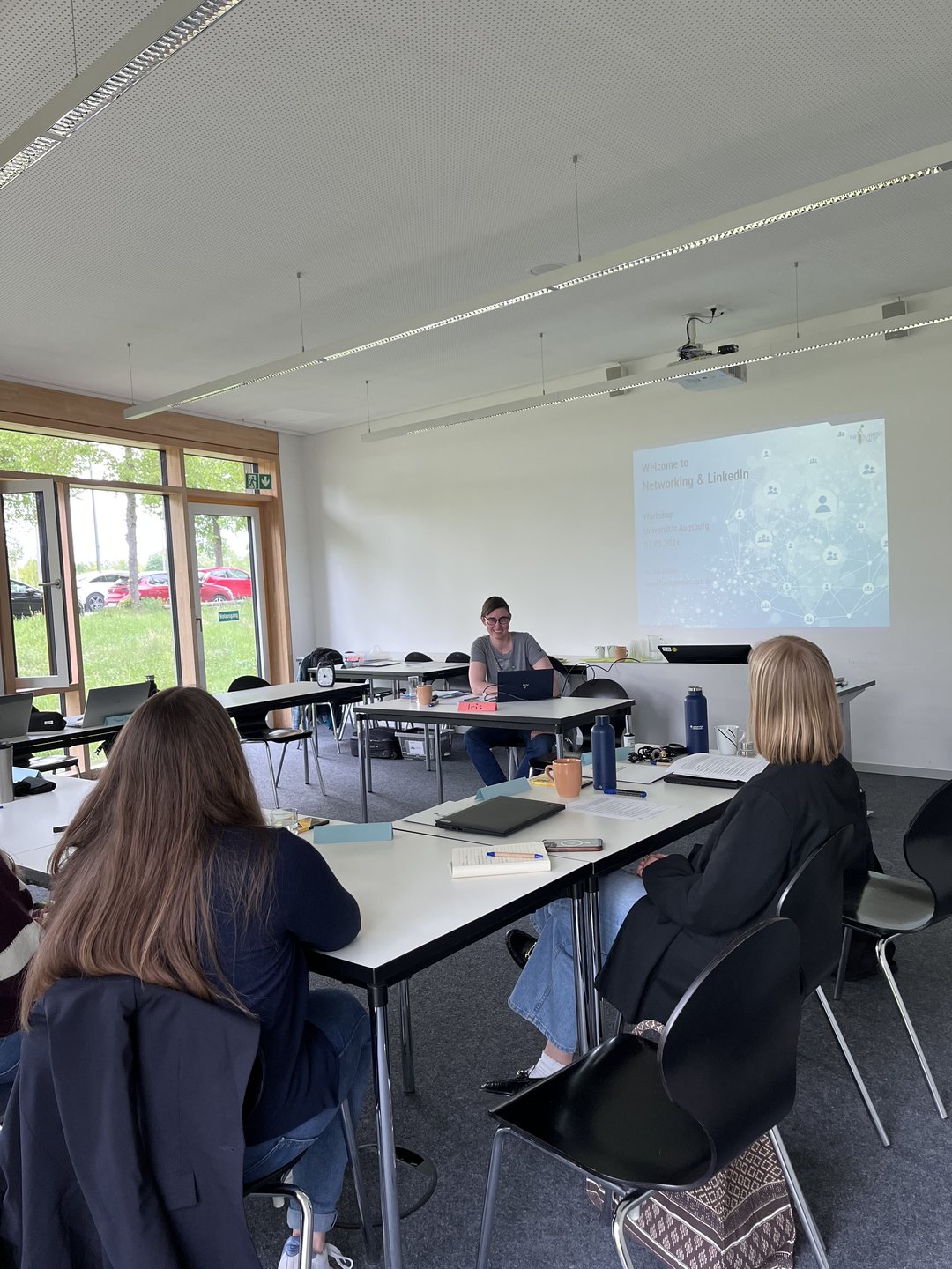 Blick in den Seminarraum mit zwei Teilnehmerinnen und der Vortragenden am Pult vorne mit der Powerpoint-Präsentation im Hintergrund.