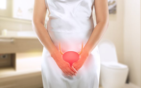 In the foreground, a woman in a white silk dress, whose head is not visible. She holds her hands crossed in front of her body. Her bladder is graphically incorporated into the image in bright red. In the background, a bright bathroom is blurred.