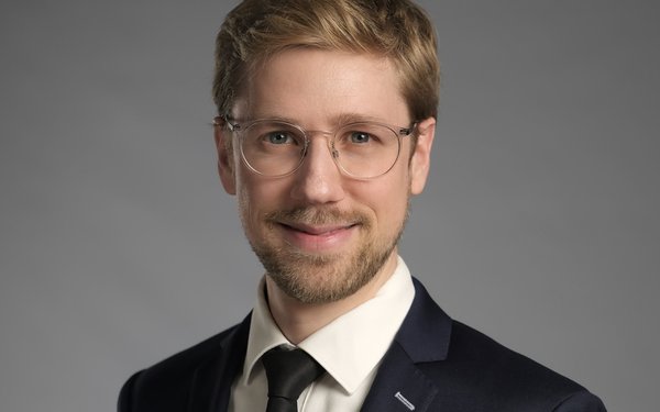 Prof. Dr. Tobias Lutzi l?chelt vor grauem Hintergrund in die Kamera. Er tr?gt eine Brille, einen schwarzen Anzug mit schwarzer Krawatte und wei?em Hemd und hat kurzes, dunkelblondes Haar.