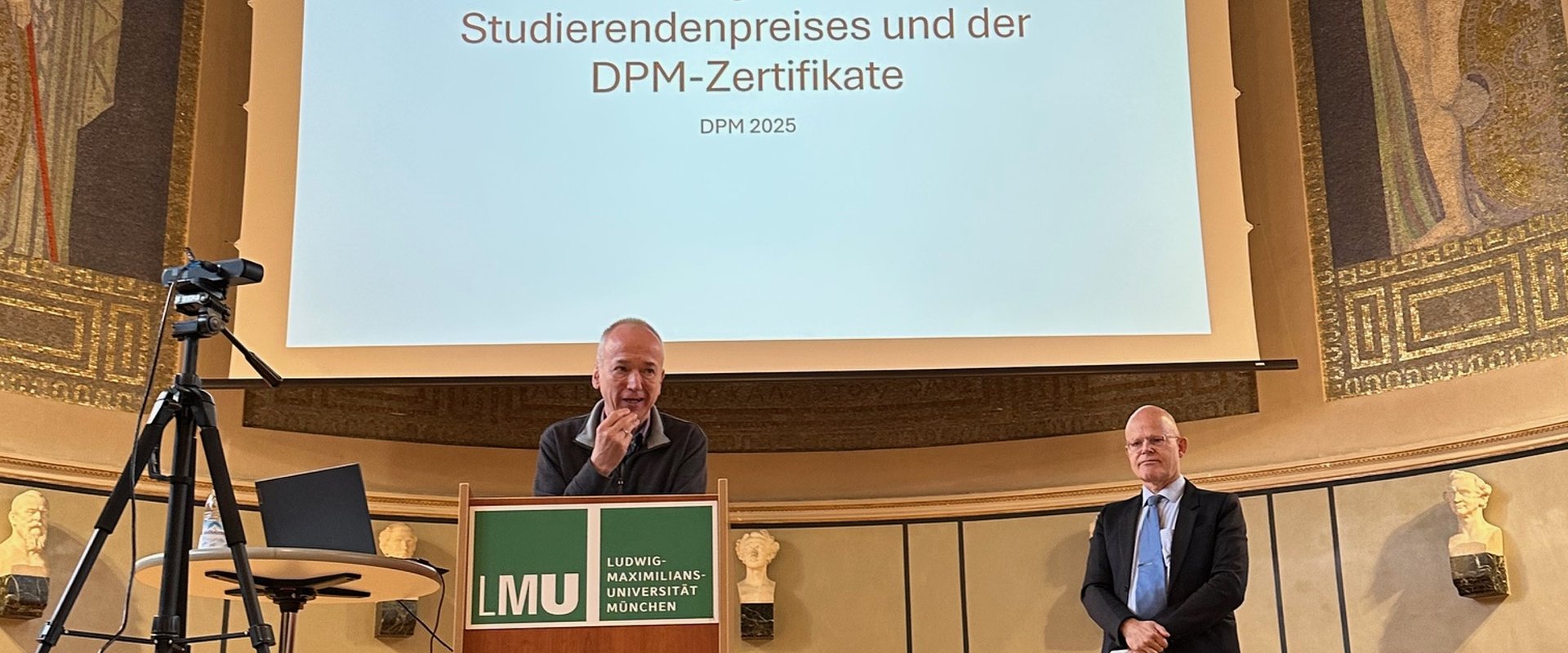 PD Dr. Karl-Heinz Selge und Prof. Dr. Dr. Elmar Gthoff stehen auf der Bhne der gro?en Aula der LMU Mnchen. Herr Selge h?lt seine Abschiedsrede und steht am Rednerpult.