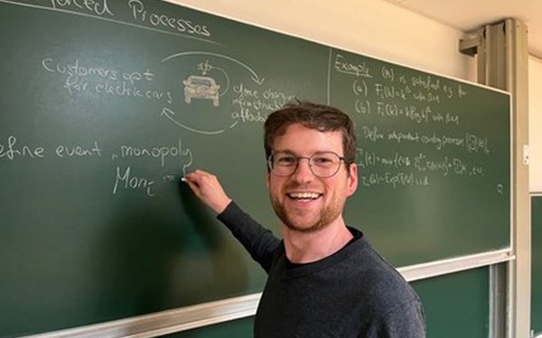 Dr. Thomas Gottfried steht vor einer Tafel, an die er mit Kreide etwas schreibt, und lächelt in die Kamera.