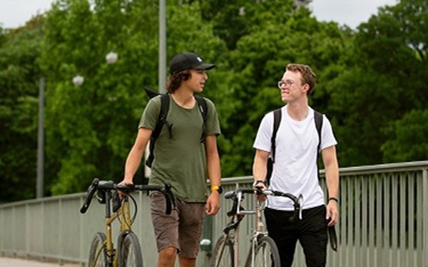 Zwei Studierende schieben ihre Fahrräder über eine Brücke und unterhalten sich. Im Hintergrund der Hotelturm