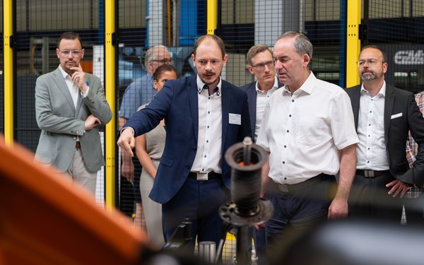 Der Wissenschaftler Prof. Markus Sause erklärt Wirtschaftsminister Hubert Aiwanger eine Forschungsanlage im KI-Produktionsnetzwerk Augsburg, während weitere Teilnehmende die Demonstration verfolgen.