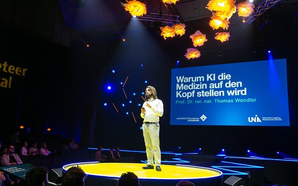 Prof. Dr. Thomas Wendler steht auf einer in blau gehaltenen Vortragsbühne in einem gelben Lichtkegel. Hinter ihm eine Leinwand mit Vortragstitel