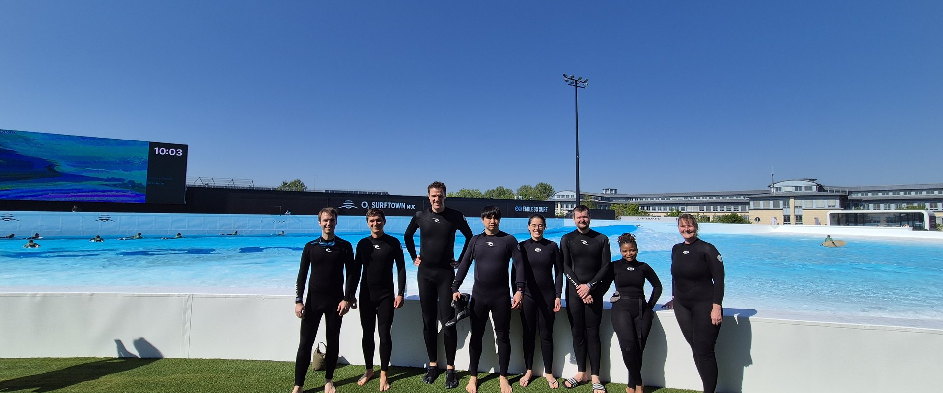 You see eight people in wet suits in front of a little wall. In the background you see the wave of the O2 surftown.