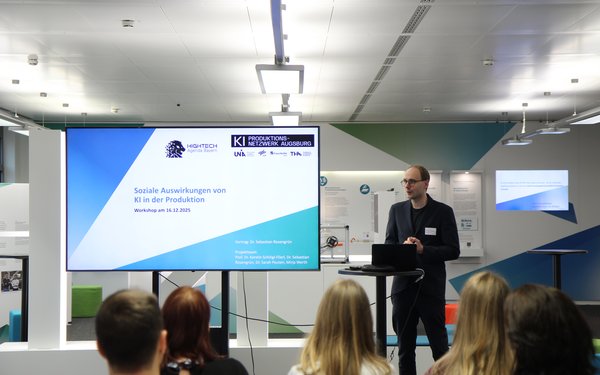 Dr. Sebastian Rosengr��n pr?sentiert seine Ergebnisse den beteiligten Unternehmen. Er steht neben einem gro?en Bildschirm, auf dem eine Pr?sentation sichtbar ist.