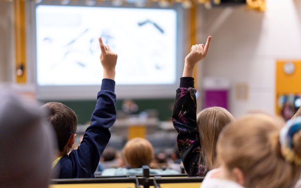 Ein Hörsaal, in den Reihen sitzen Kinder. Im Vordergrund melden sich ein Junge und ein Mädchen.