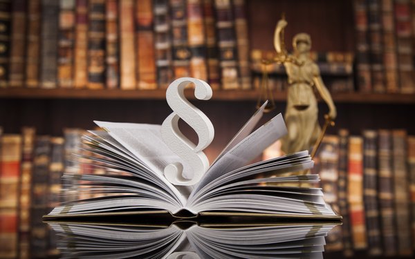 Ein aufgeschlagenes Buch und eine Statue der Justitia im Hintergrund deuten einen juristischen Zusammenhang an.