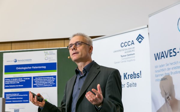 Prof. Dr. Martin Trepel steht vor Roll-ups mit Krebsthematik, gestikuliert und h?lt einen Vortrag.