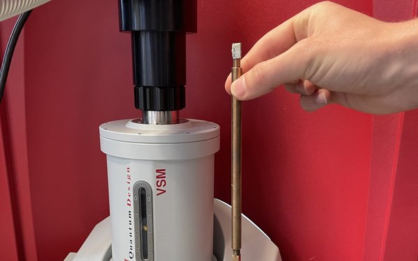 Eine Hand hält einen messingfarbenen Stab neben einem kompliziert aussehenden Gerät, dem Magnetometer.