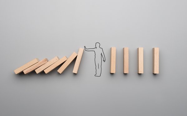 Eine Reihe von Dominosteinen. Die erste Hälfte ist umgefallen, zwischen dieser und der zweiten Hälfte steht ein gezeichneter Mann und streckt den Arm nach vorne. Hierdurch schützt er die weiteren Steine vom fallen.