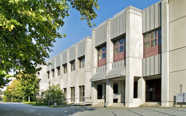 Blick von schräg-vorne auf den Eingangsbereich eines brutalistischen grauen Beton-Gebäudes. Auf der linken Bildseite grüne Bäume und Sträucher.