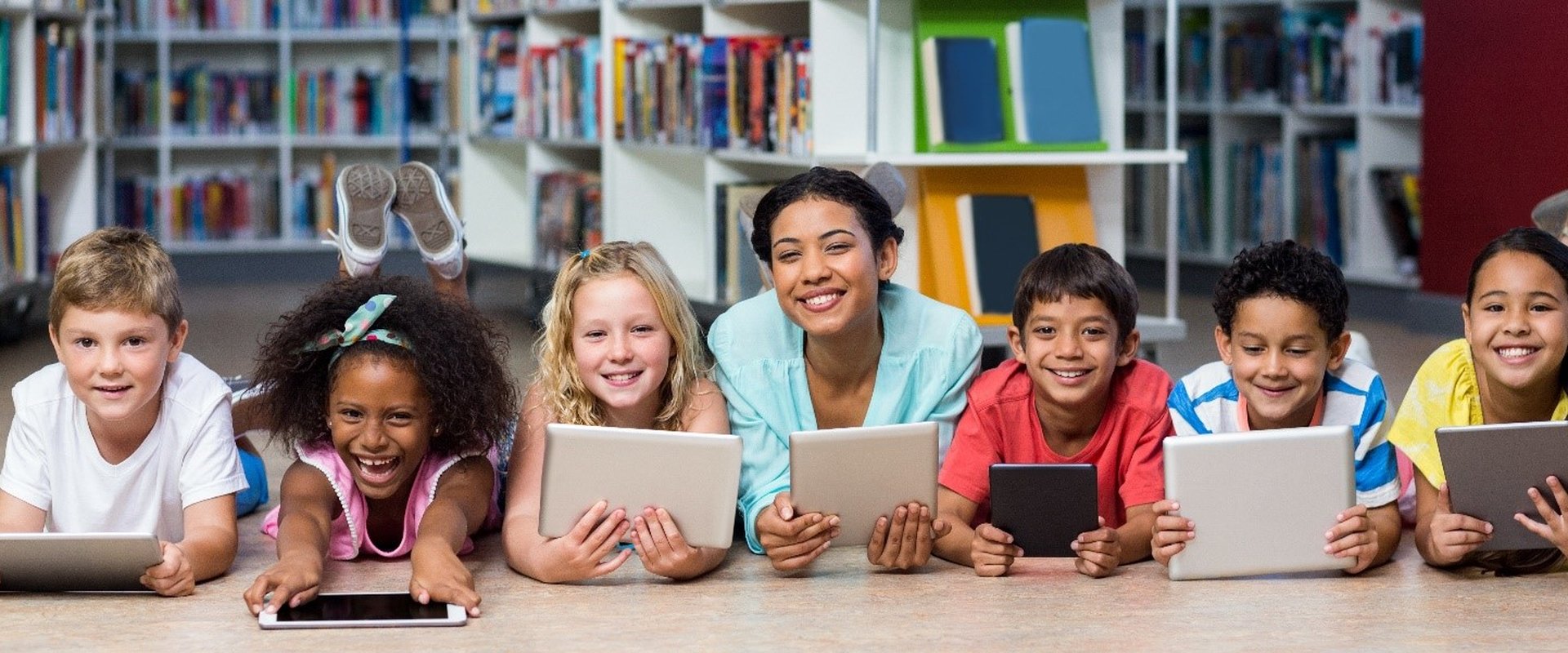 Eine Reihe von 6 Kindern und einer Lehrkraft. Sie liegen auf dem Boden mit Blick nach vorne, die Lehrerin in der Mitte. Jede/jeder hält ein iPad vor sich und blickt gut gelaunt.