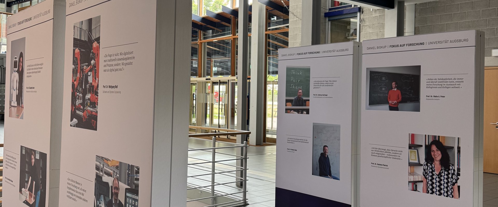 In einer hellen Universitätslobby stehen mehrere hohe Ausstellungstafeln mit Fotos und Zitaten von Forschern der Universität Augsburg.