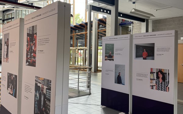 In einer hellen Universitätslobby stehen mehrere hohe Ausstellungstafeln mit Fotos und Zitaten von Forschern der Universität Augsburg.