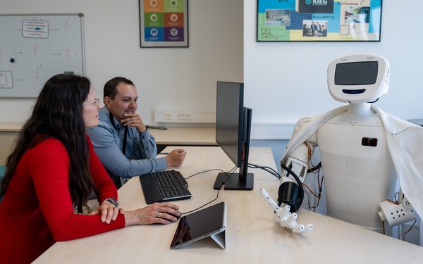 Zwei Menschen mit Tablet-Computern sitzen einem Roboter gegenüber.