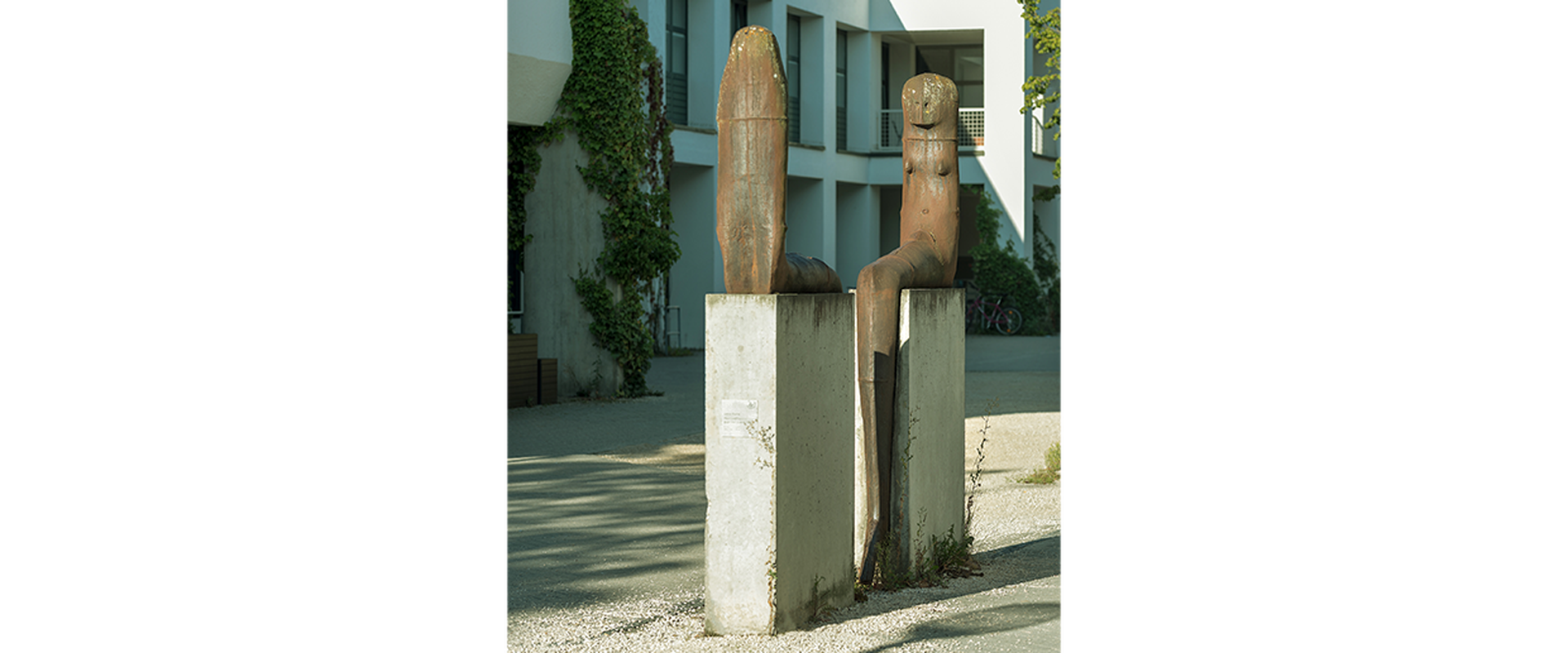 Auf dem Bild sieht man das Kunstwerk von Lothar Fischer, bei dem sich zwei Skulpturen gegenübersitzen auf Steinen.