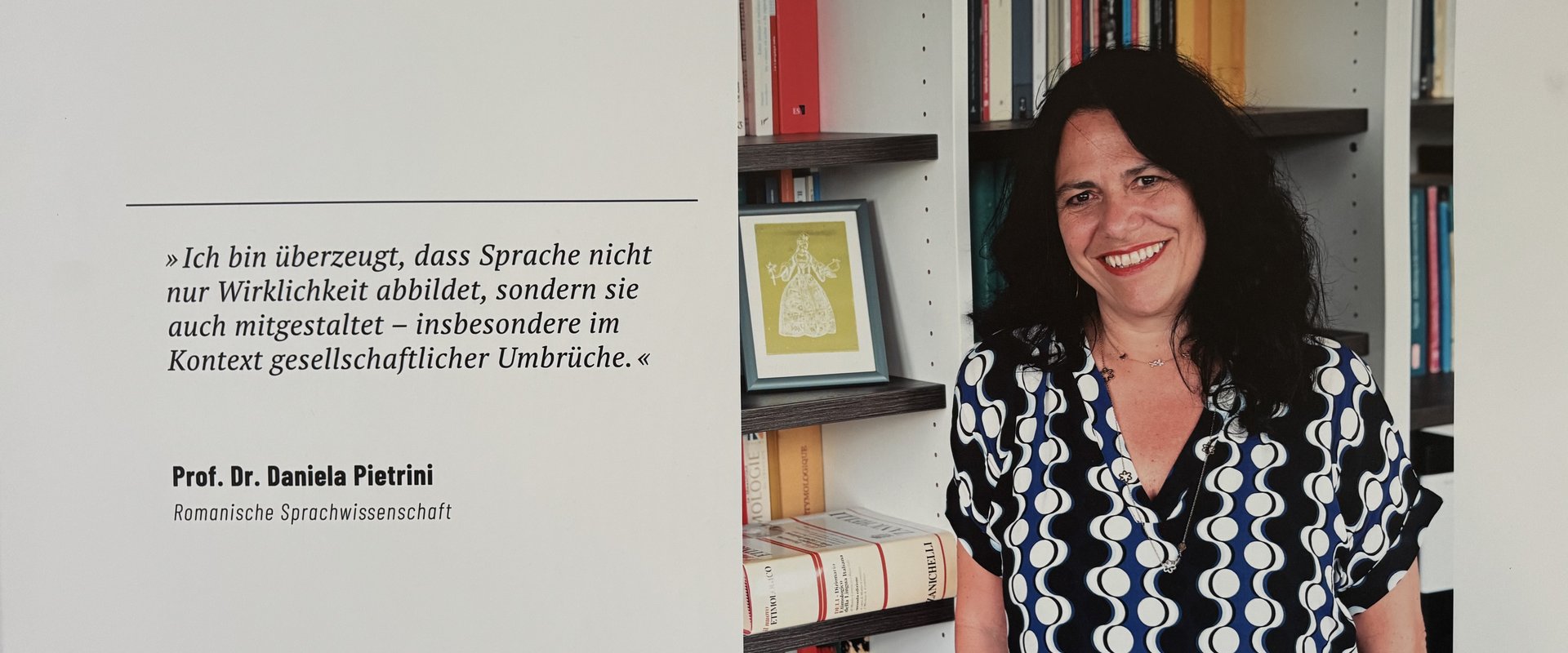 Eine Ausstellungstafel zeigt das Porträt einer lächelnden Wissenschaftlerin vor einem Bücherregal. Links daneben steht ein Zitat über Sprache als gestaltende Kraft gesellschaftlicher Wirklichkeit sowie Name und Fachgebiet der Professorin.