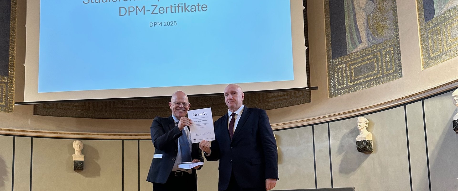 Prof. Dr. Dr. Elmar Gthoff berreicht den Studierendenpreis an Declan O'Keeffe. Beide stehen auf der Bhne der Gro?en Aula der LMU Mnchen.
