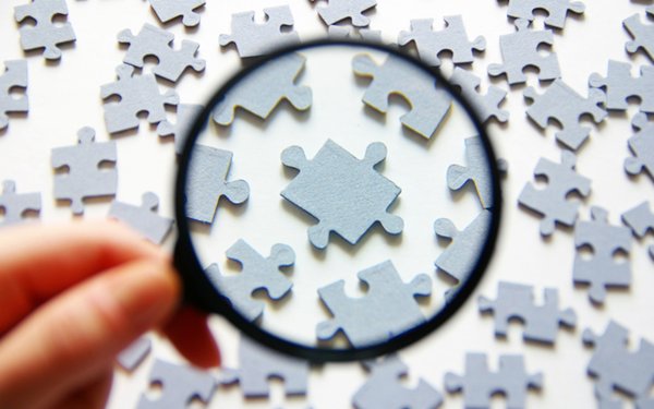 Symbolbild: Eine Hand hält eine Lupe über graue Puzzelstücke, die auf einer weißen Fläche liegen.