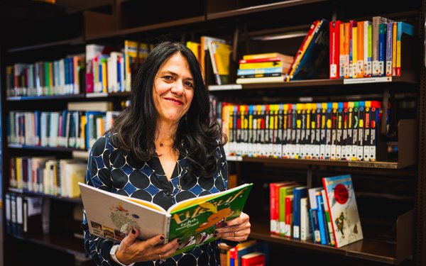 Prof. Dr. Daniela Pietrini steht vor einem Bücherregal, hat ein aufgeschlagenes Comicbuch in der Hand und lächelt in die Kamera.