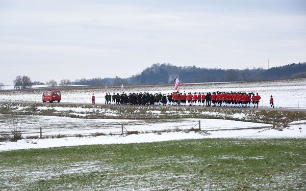 Eine Gruppe von rot-schwarz gekleideten Dinkelscherbener Schäfflern geht auf einer Straße, die Landschaft ist Schnee bedeckt