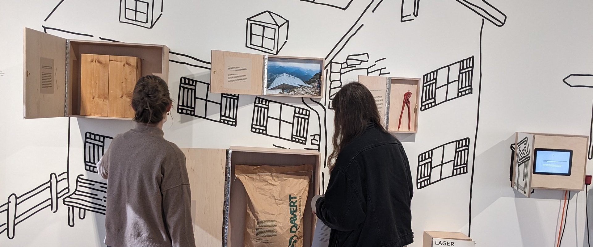 Zwei Personen stehen vor einer Museumswand, wo ein Haus abgebildet ist und darauf sind K?sten mit Ausstellungsst��cken