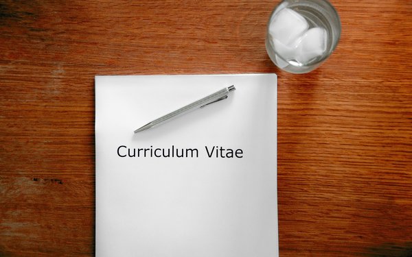 Das Bild zeigt ein auf einem Holztisch liegendes Papier, auf dem "Curriculum Vitae" steht