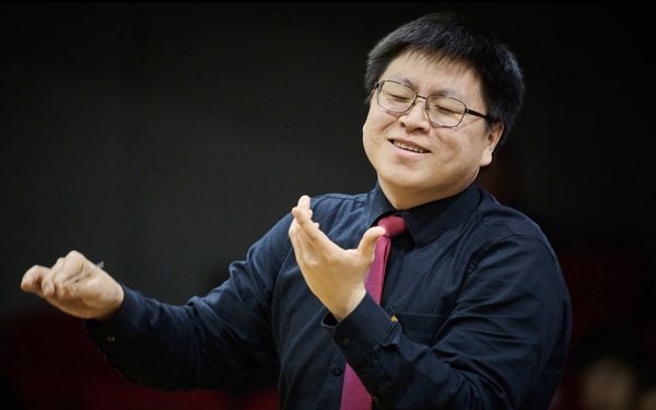 Jen-Jen Liu vor schwarzem Hintergrund, in dunkelblauem Oberteil und mit bordeaux-roter Krawatte. Er tr?gt eine Brille, hat die Augen geschlossen und macht ausladende Dirigierbewegungen mit den Armen