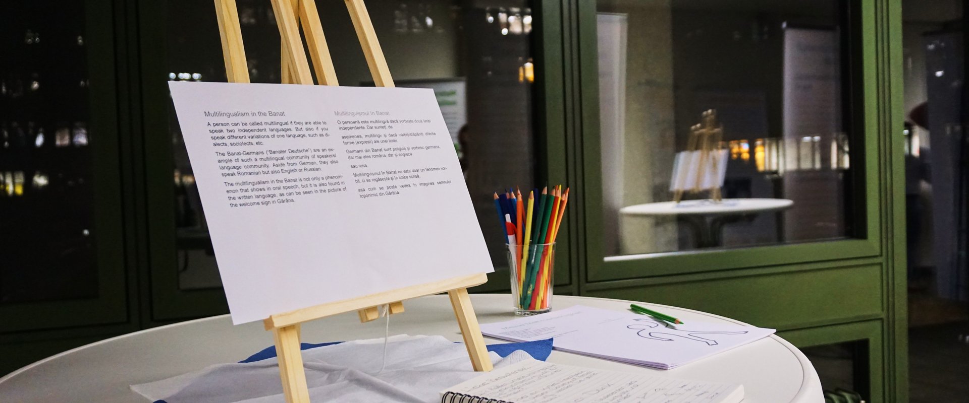 Staffelei mit englischer und rumänischer Zusammenfassung eines Roll-Up-Banners, Gästebuch und Farbstiften.