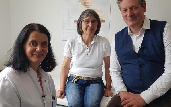 Ein Behandlungszimmer. Links sitzt die Professoring, in der Mitte sitzt auf der Liege eine ältere Patientin, rechts von dieser sitzt Herr Hirschhausen. Alle lächeln.