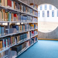 Links vom Gang ein regal mit vielen Büchern, rechts vom Gang Glastüren, geradeaus ein rundes Fenster