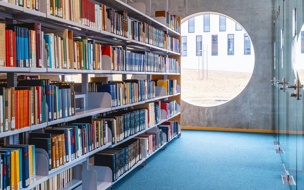 Links vom Gang ein regal mit vielen Büchern, rechts vom Gang Glastüren, geradeaus ein rundes Fenster