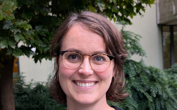 Dr. Lisa Kolb l?chelt in die Kamera. Sie tr?gt eine Brille und hat hellbraunes, kinnlanges Haar. Im Hintergrund ein Baum und der Teil eines Uni-Geb?udes.