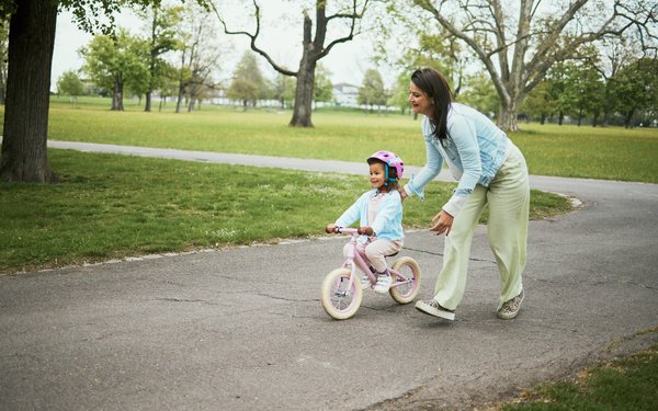 Ein kleines M?dchen f?hrt in einer Parklandschaft mit einem rosaroten Kinderfahrrrad. Es tr?gt einen rosaroten Helm und ein hellblaues Oberteil. Eine Frau l?uft neben dem Fahrrad und h?lt es zur Sicherheit fest.