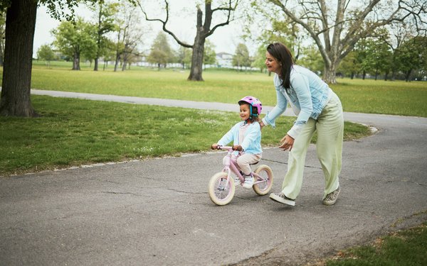 Ein kleines M?dchen f?hrt in einer Parklandschaft mit einem rosaroten Kinderfahrrrad. Es tr?gt einen rosaroten Helm und ein hellblaues Oberteil. Eine Frau l?uft neben dem Fahrrad und h?lt es zur Sicherheit fest.
