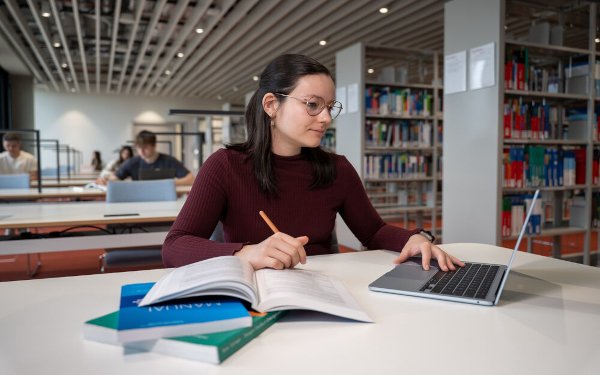 Eine junge Frau mit dunkelrotem Pullover, Brille und halblangem dunklem Haar sitzt in einer Bücherei am Tisch und lernt. Auf dem Tisch liegen mehrere aufgeschlagene Bücher und ein Laptop, an dem die Frau arbeitet.